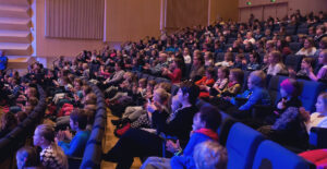 Paljon ihmisiä istumassa penkeillä Rauma-salissa katsoen esitystä.