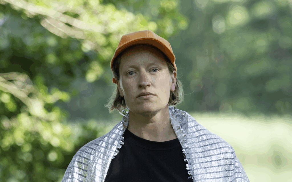 Taiteilija Maija-Annikki Savolanen kuvattuna rinnasta ylöspäin ja katsoo suoraan kameraan vakavana Päässään hänellä on oranssi lippalakki ja päällään musta oaita ja hartioilla valkoinen huivi. Taustalla kuvassa näkyy vihreää luontoa.