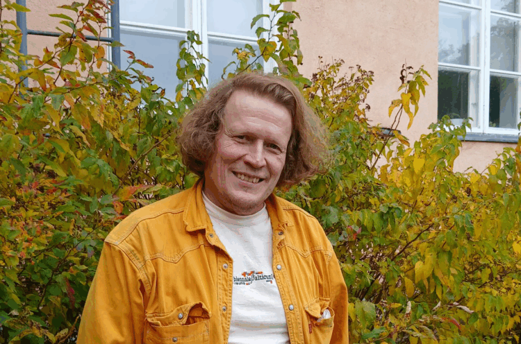 The artist looks at the camera and smiles. He is wearing a yellow shirt, and a pink museum building can be seen behind him.