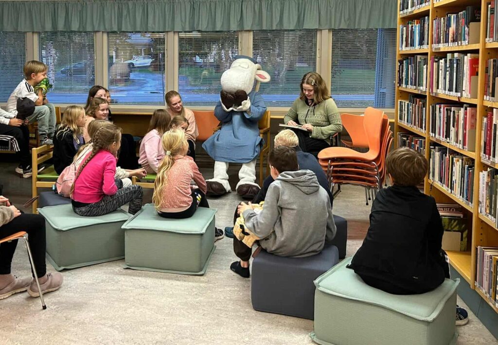 Kuvassa lapsia ja Herra Hakkarainen kirjastossa. Lapsille luetaan kirjaa.