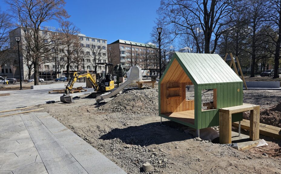 Keskuspuiston rakennustyömaalla asennetaan leikkivälineitä. Kuvassa pieni mökki.