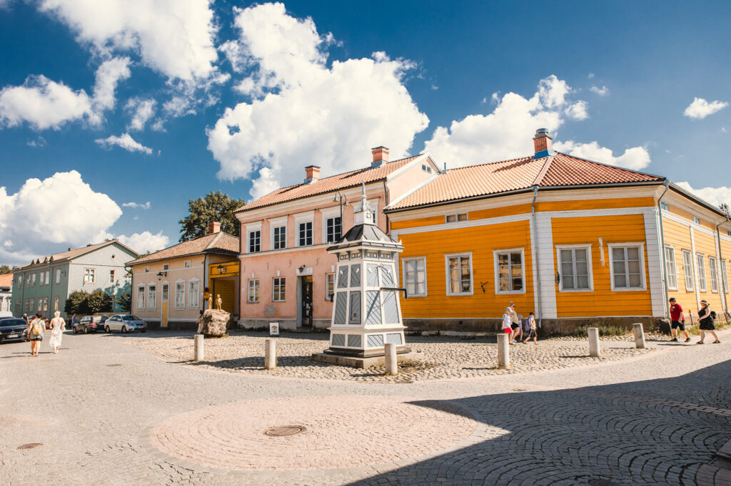 Maisema taidemuseon julkisivusta. Aurinko paistaa ja taivaalla valkoisia pilviä. Rivissä vanhoja värikkäitä taloja ja talojen edessä vanha sininen puukaivo.