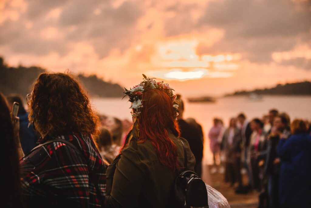 A woman wearing a flower crown at a festival, looking towards the sunset. People can be seen in the background.