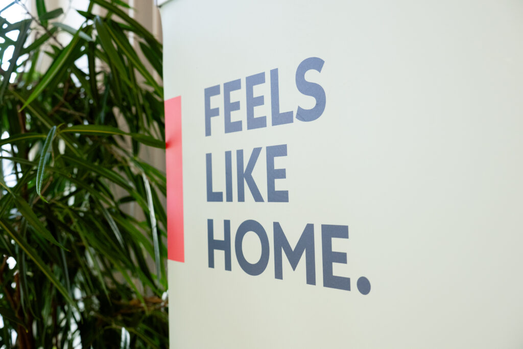 The International House Rauma slogan “Feels Like Home” is displayed on a wall. A red line is painted next to the slogan, and a plant stands beside the wall.