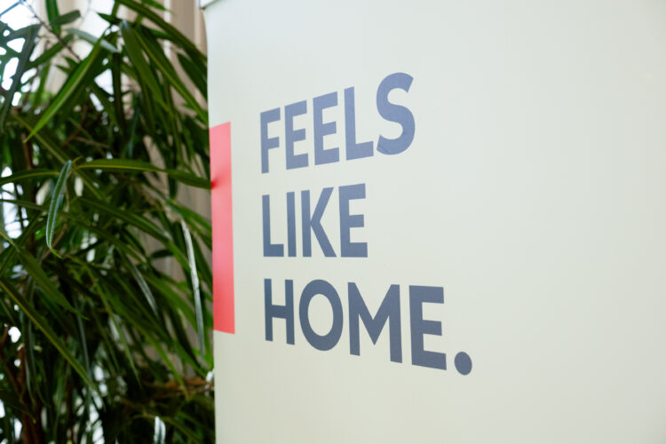 The International House Rauma slogan “Feels Like Home” is displayed on a wall. A red line is painted next to the slogan, and a plant stands beside the wall.