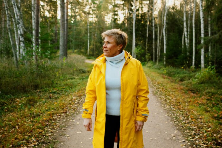 Kuvassa lyhythiuksinen nainen, jolla on keltainen ulkoilutakki päällä. Nainen on kävelemässä polulla. Taustalla metsää.