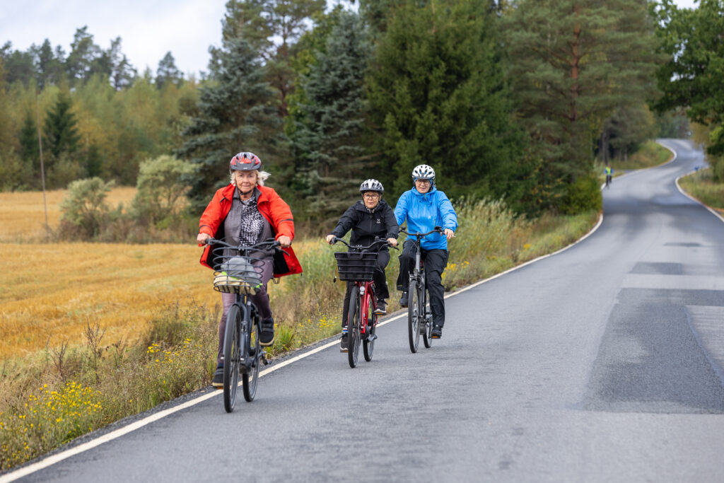 Kolme naista pyöräilemässä autotien varrella. Taustalla metsää ja peltoa.