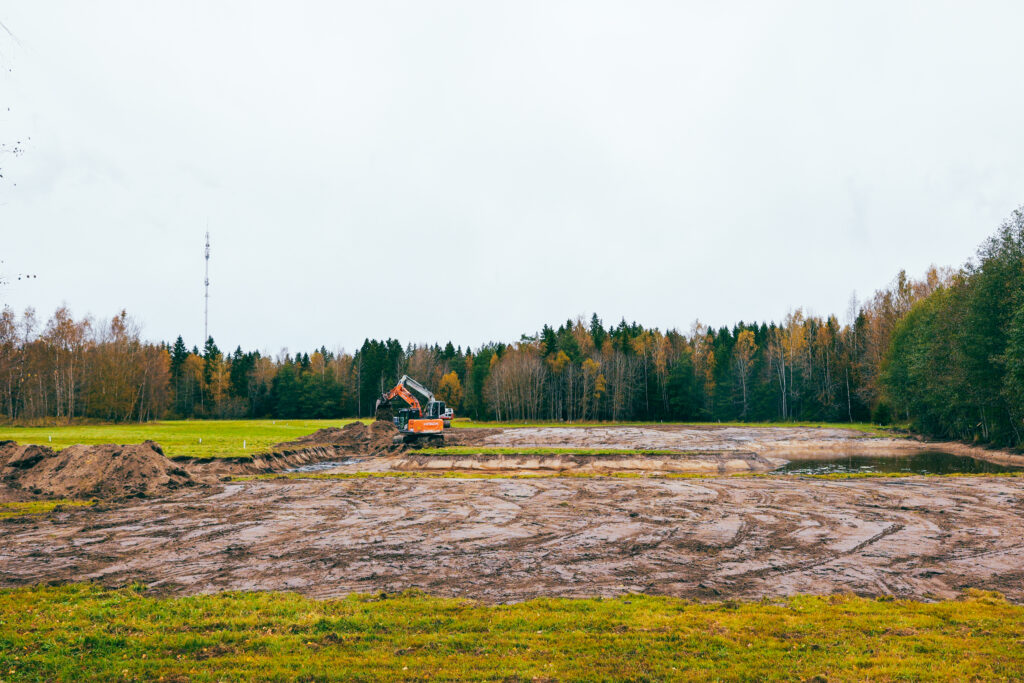 Kosteikon rakennustyöt Pohjoiskehällä. Kuvassa kaivinkone työskentelee osittain raivatulla peltoalueella, jota ympäröi metsä. Alueelle rakennetaan kosteikko Rauman DiscGolf-radan yhteyteen osana vesiensuojelutyötä.